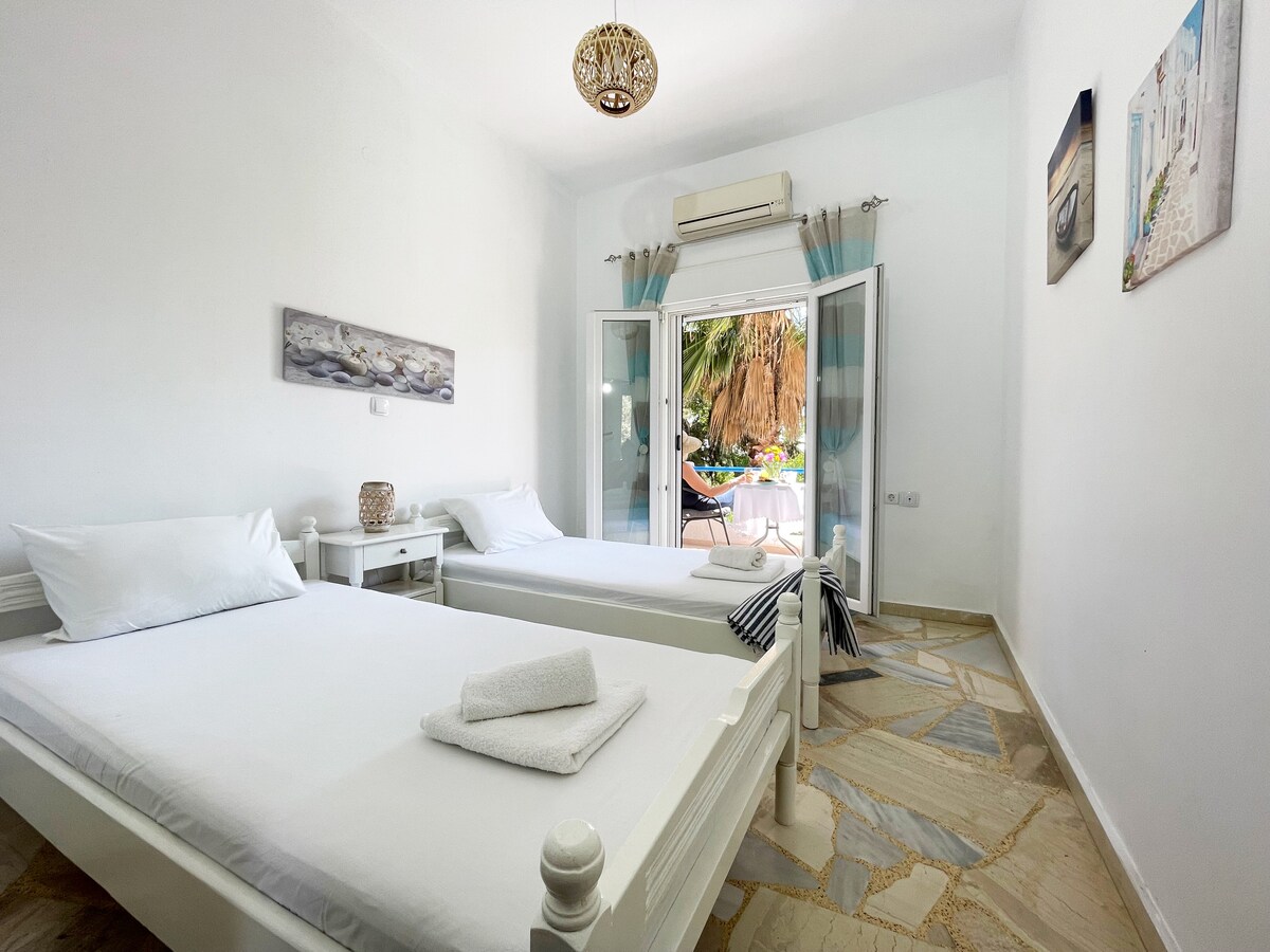A bright and airy room features two twin beds with white linens, strategically placed near a large window. Light-colored walls create an open atmosphere, and decorative artwork adorns the walls. A glimpse of outdoor greenery can be seen through the open French doors.