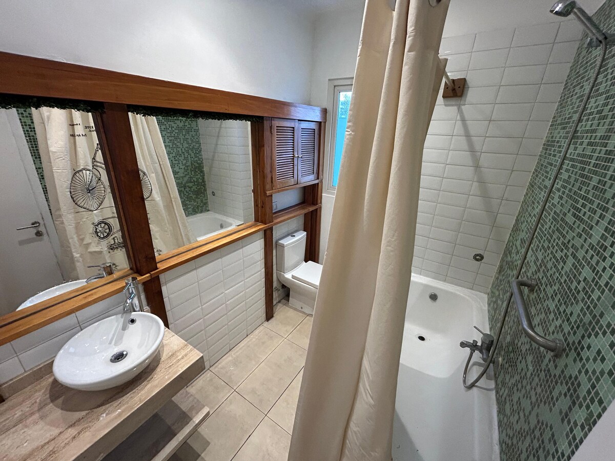A well-appointed bathroom features a bathtub alongside a shower area, with a white basin placed on a natural wood counter. A large mirror reflects the space, while green tile accents add a calming touch. Natural light enters through a nearby window.