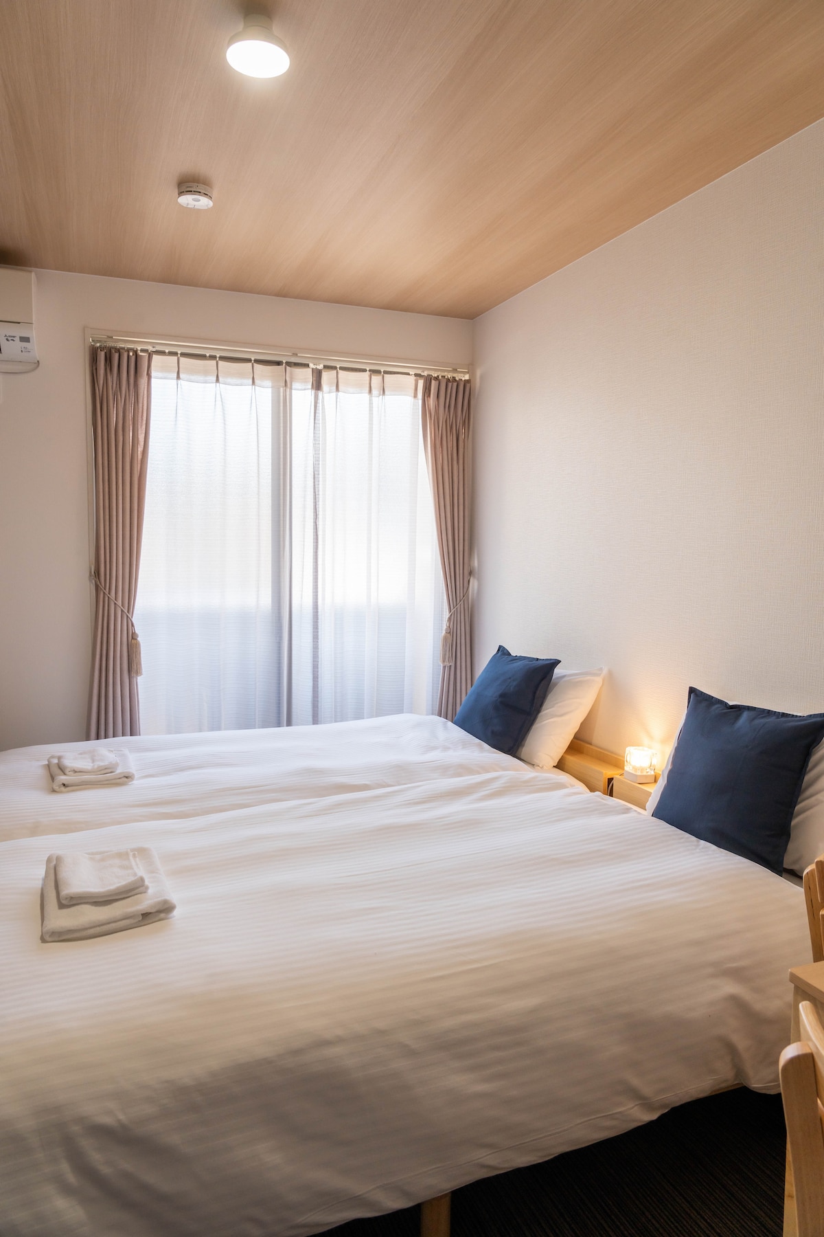 A cozy bedroom features two spacious beds with crisp white linens and matching dark cushions. Natural light filters through sheer curtains, illuminating the room's warm wood accents and subtle decor. A soft glow emanates from a small bedside lamp, adding to the serene atmosphere.