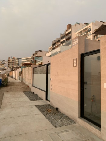 The exterior view showcases a modern building design with smooth, neutral-toned walls and large glass doors. A paved pathway runs alongside, bordered by small gravel areas. Nearby structures provide a contemporary architectural background, complementing the clean lines of the property.