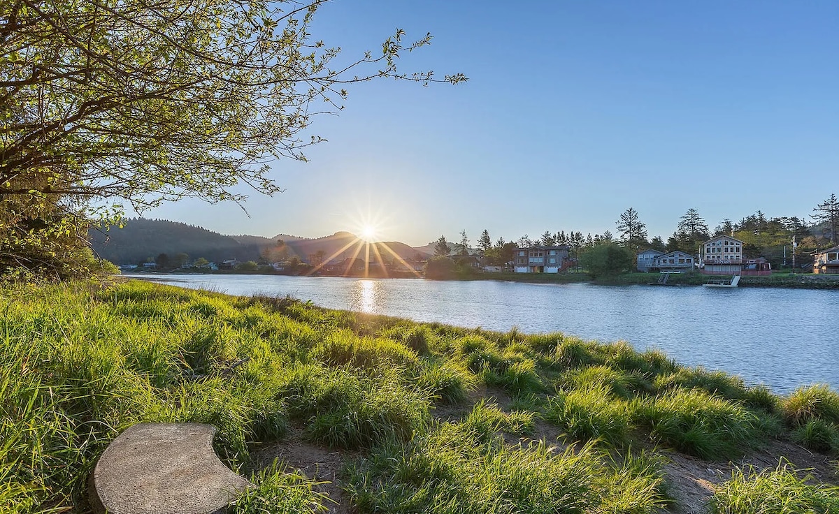 A serene riverside view captures the sun rising over the water, with lush green grass in the foreground. The reflection of the sunlight dances on the surface, while trees and colorful buildings are visible on the opposite bank, enhancing the tranquil setting.