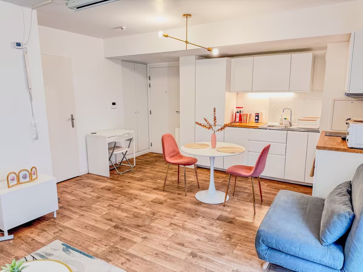A bright living area features a comfortable sofa and a round dining table surrounded by pink chairs. The open kitchen is neatly arranged with modern appliances and storage. Light wood flooring adds warmth to the space, complemented by ample natural light from nearby windows.
