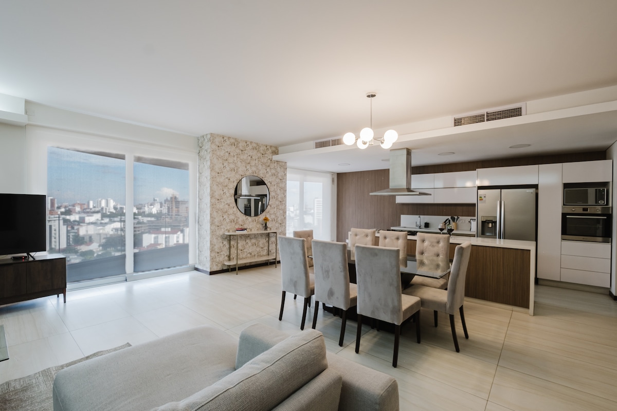 An open-plan living and dining area features a modern kitchen with sleek cabinetry and stainless steel appliances. A dining table with upholstered chairs accommodates several guests. Large windows allow natural light to fill the space, offering a view of the city skyline.