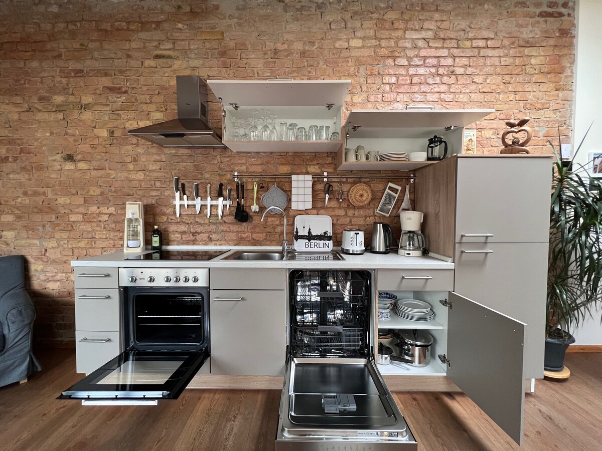 A modern kitchen showcases an assortment of appliances, including an oven, dishwasher, and coffee machine. Open shelves display glassware, while utensils are neatly arranged on a rack. The warm brick wall adds character, completing the functional design that encourages cooking and dining.