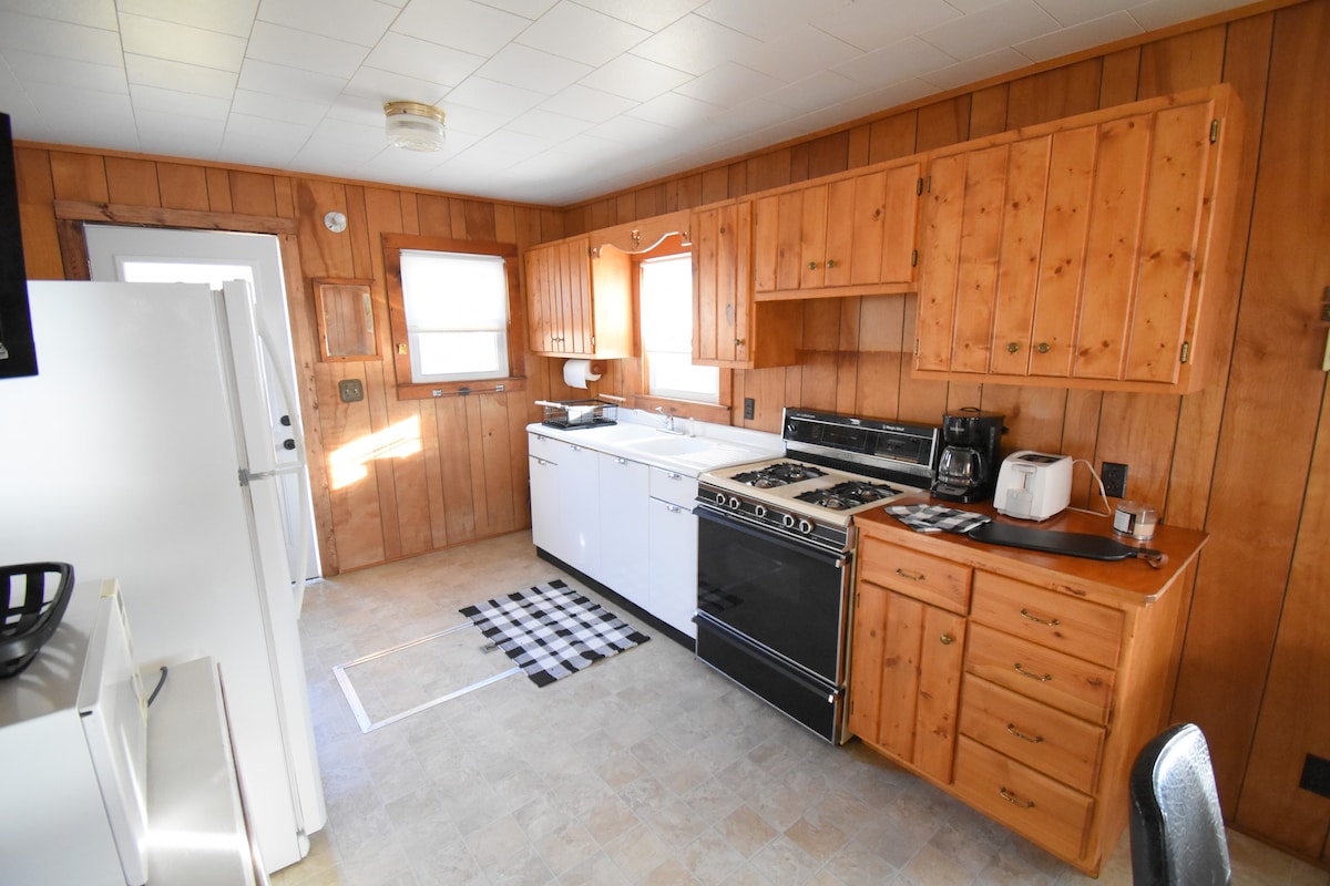 The kitchen features wooden paneling, a gas stove, and a wide counter space. A refrigerator stands to the left, while a white dishwasher is integrated into the cabinetry. A black and white checkered rug lies on the floor, adding a touch of contrast.