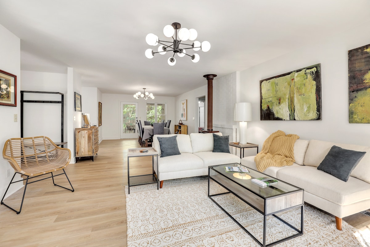 The living room features a spacious layout with comfortable seating arrangements. A large, contemporary coffee table is centered on a soft area rug. Natural light filters through the windows, highlighting the art pieces on the walls and creating a bright, inviting space.