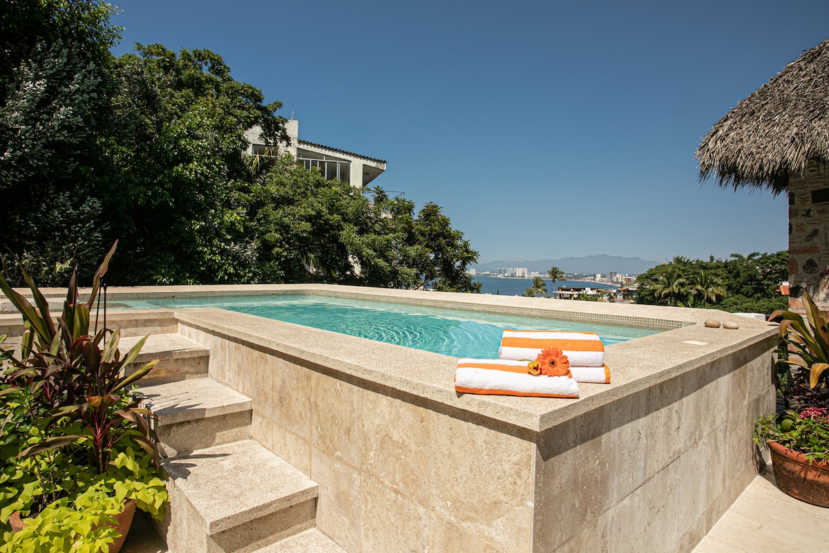 A serene swimming pool is surrounded by landscaped greenery, featuring steps that lead into the clear water. Soft orange towels are neatly arranged on the edge, while a thatched roof shade provides a relaxed atmosphere. Views of the distant hills and ocean are visible.