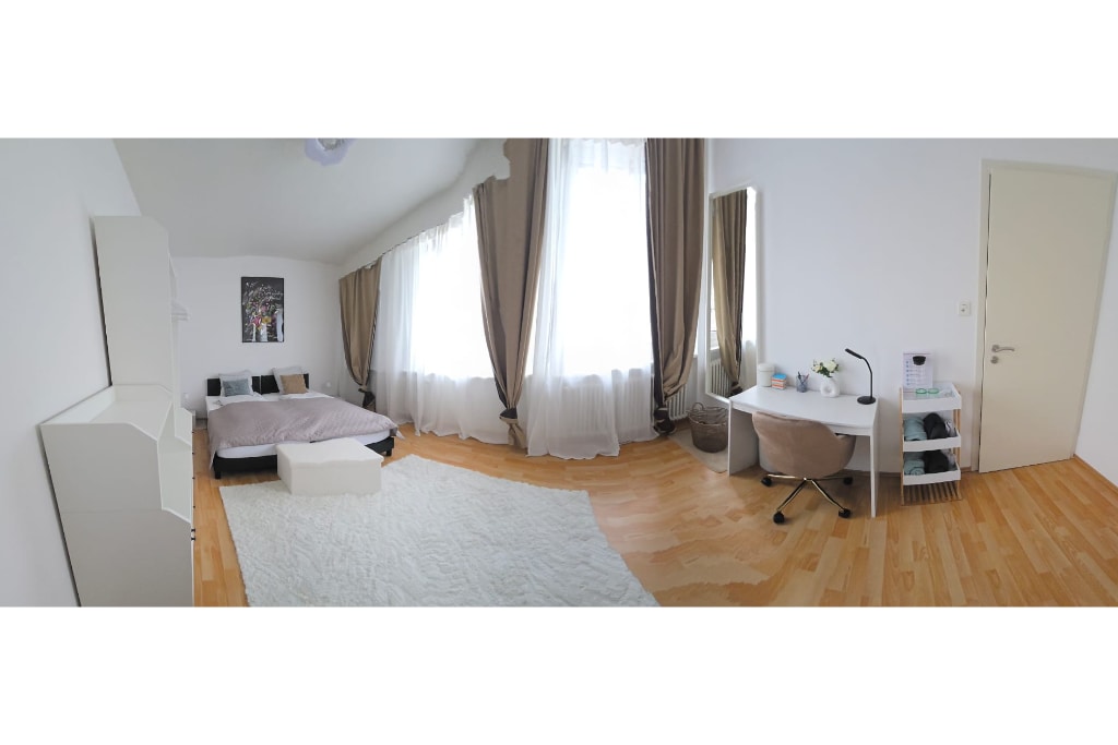 A spacious room features a large bed with soft bedding and a fluffy area rug underneath. Natural light filters through tall windows adorned with light curtains. A desk with a chair and a mirror are positioned against the wall, contributing to the room's bright atmosphere.
