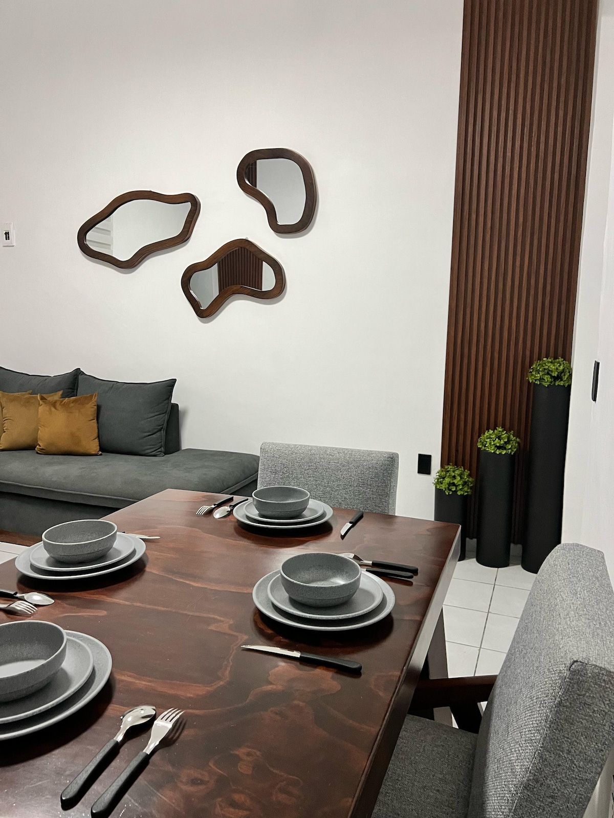 A modern dining area features a dark wooden table set with grey dishware and stainless steel utensils. A comfortable sofa with decorative cushions is arranged nearby. Organic-shaped mirrors adorn the wall, complemented by vertical planters with greenery.