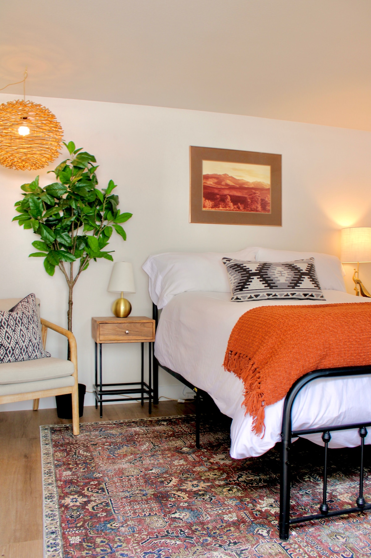 A cozy sleeping area features a spacious bed dressed with a bright orange throw and decorated with a patterned pillow. A bedside table with a lamp sits beside the bed. A green potted plant and a small chair enhance the inviting space.