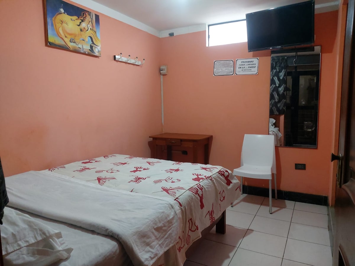 A simple room features two beds with white linens, accented by a red patterned blanket. A small wooden desk and a white chair are positioned against the wall. A television is mounted above the desk, and a window allows natural light to enter the space.
