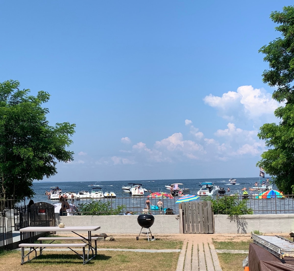 A view of a calm lake is captured, with several boats anchored nearby. Colorful umbrellas dot the shoreline, while a grassy area with picnic tables and a charcoal grill is visible in the foreground. Trees frame the scene, contributing to the relaxed atmosphere.