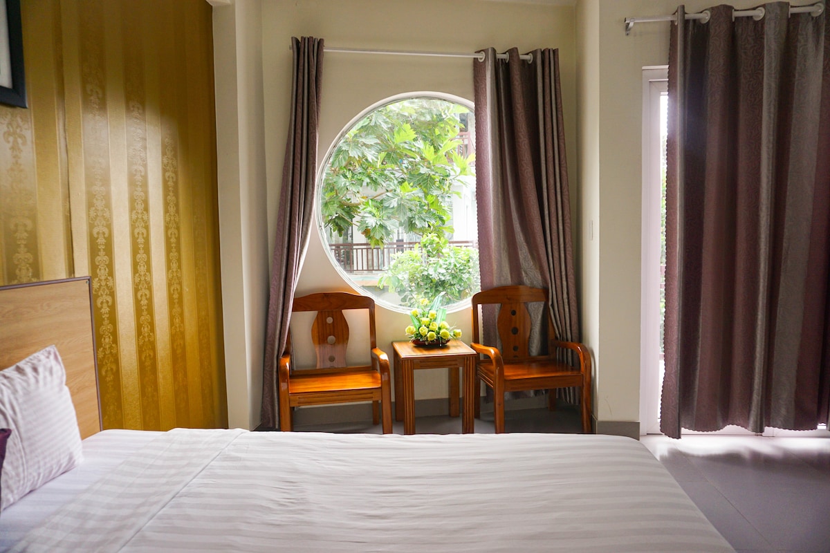 A spacious room features a large bed with crisp linens and a rounded window offering views of the greenery outside. Two wooden chairs are positioned beside a small table adorned with a decorative plant. Elegant curtains frame the window, enhancing the inviting ambiance.