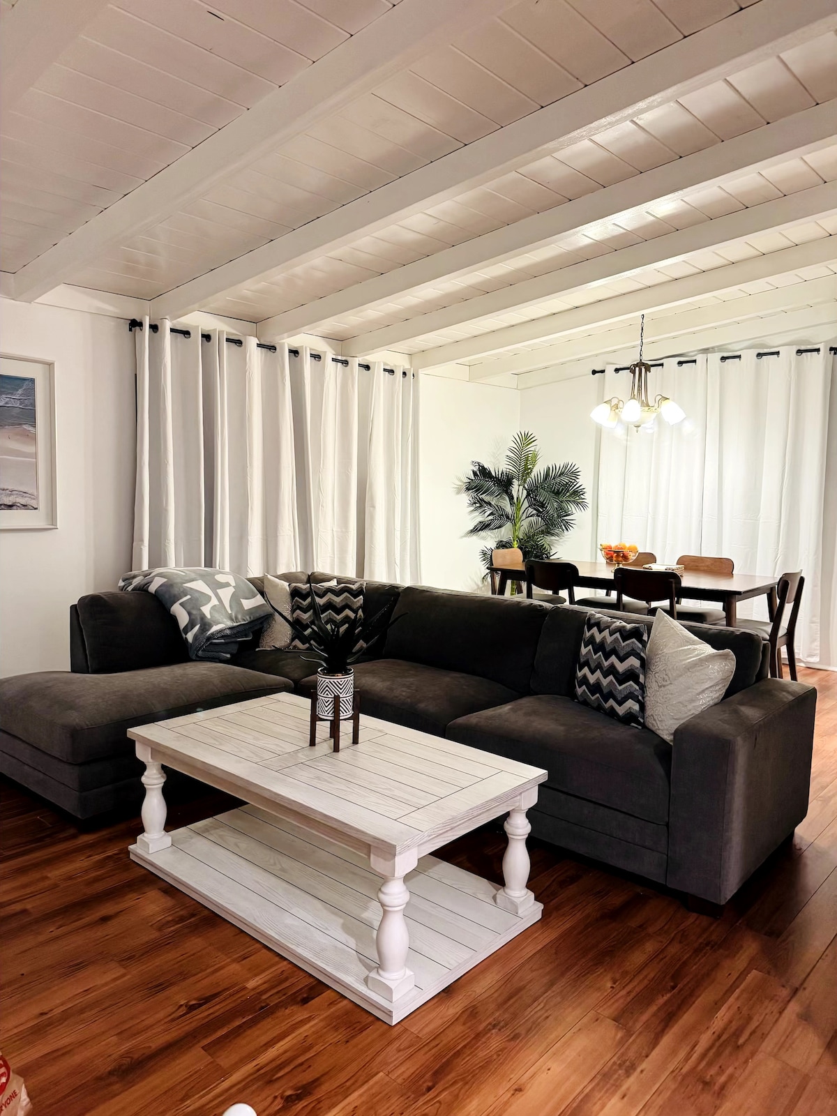A spacious living area features a dark sectional sofa adorned with patterned accent pillows. A white coffee table rests in front, with a decorative centerpiece. In the background, a dining area with a wooden table and chairs is illuminated by a pendant light, surrounded by sheer curtains.