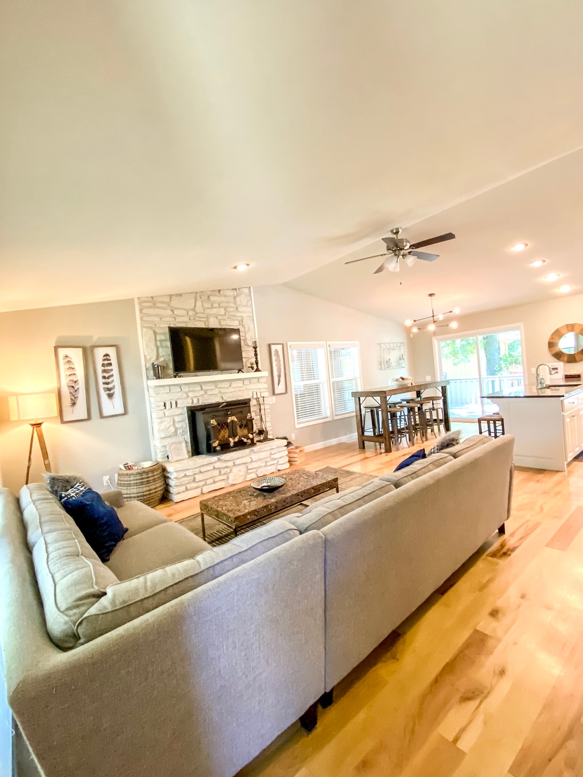A light-filled living space features a cozy sectional sofa facing a stone fireplace, complete with a TV mounted above. A hardwood floor extends through the area, leading to a dining space with tall chairs and a table in view. Large windows reveal the outdoors.