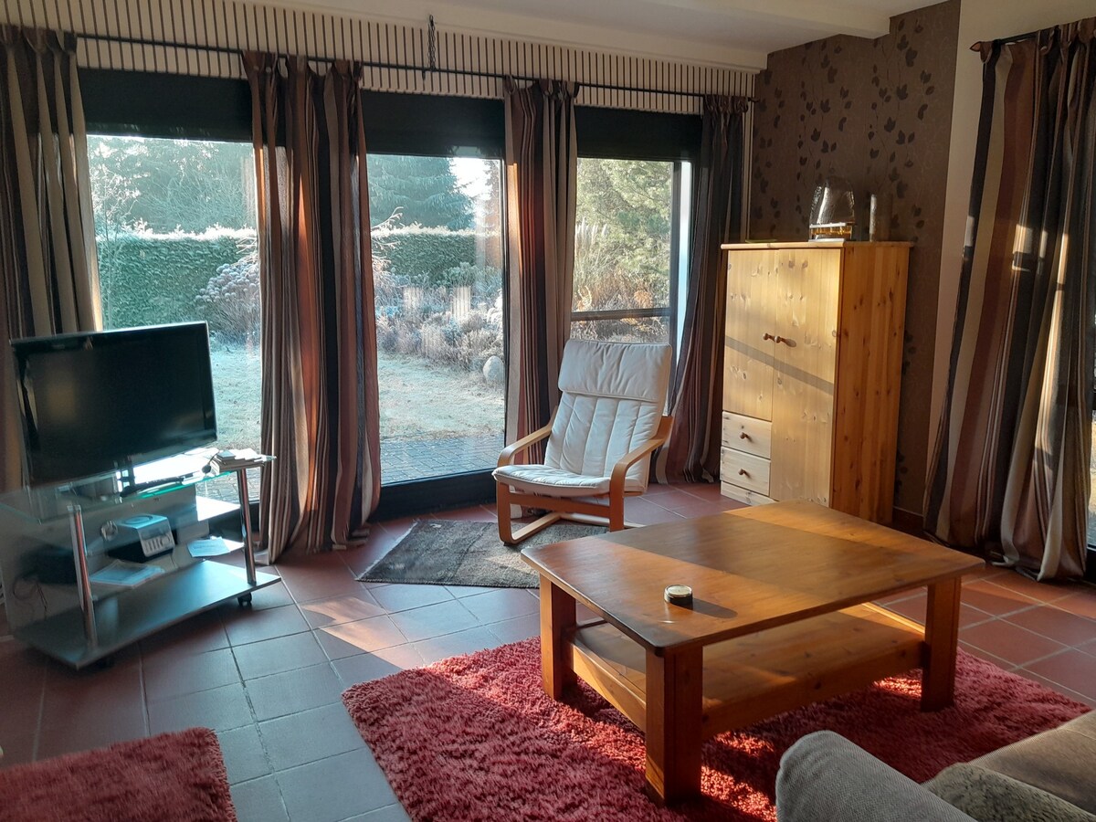 A spacious living room is highlighted by large glass doors allowing natural light to fill the area. A comfortable armchair and a wooden coffee table are present, with a modern TV setup on a glass stand. Soft rugs and elegantly draped curtains enhance the cozy setting.