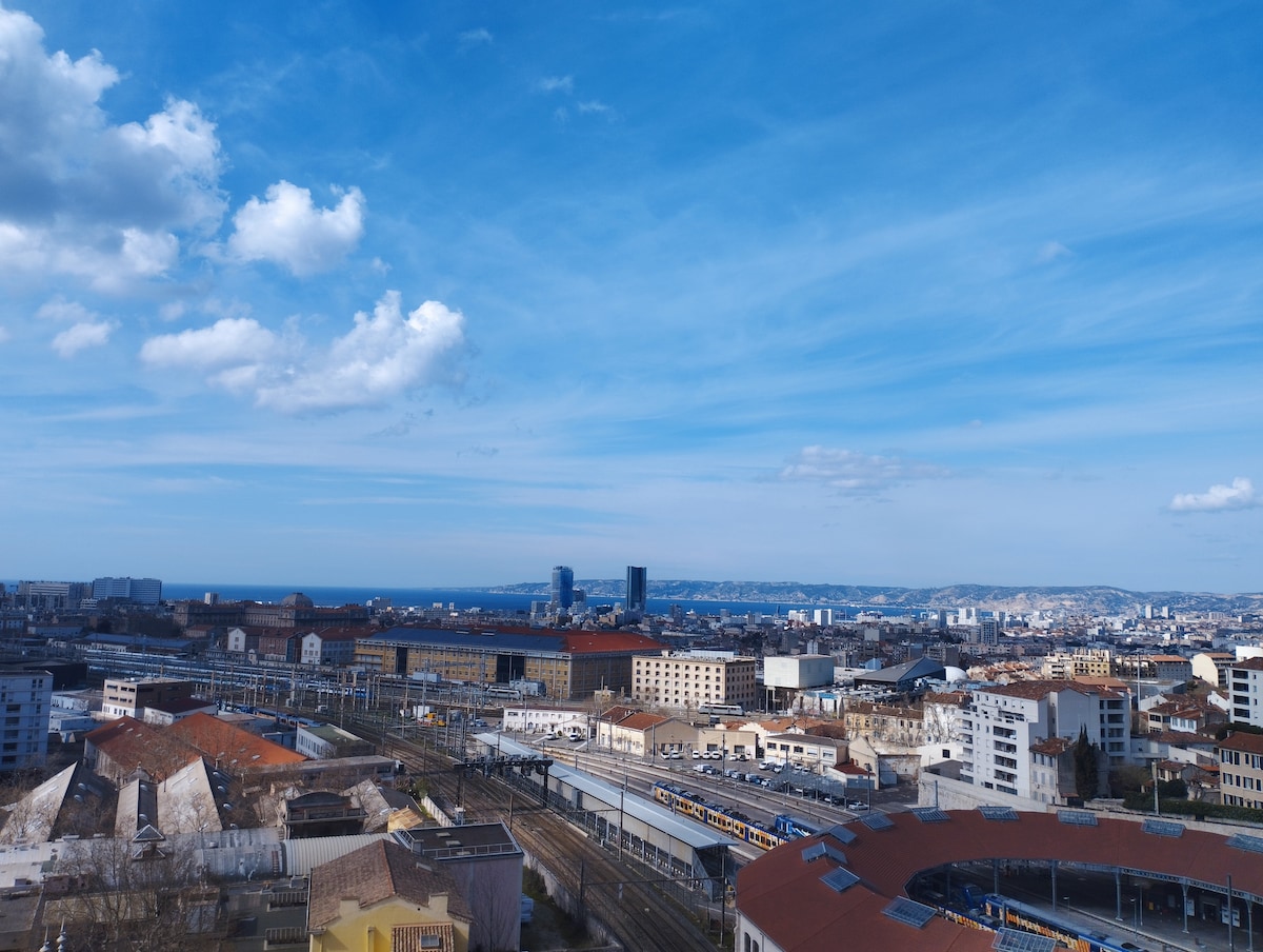 A panoramic view of Marseille is showcased, featuring the cityscape with a distant view of the sea and hills. Notable buildings and structures are present, with trains visible on tracks below. The sky is partly cloudy, enhancing the clarity of the landscape.