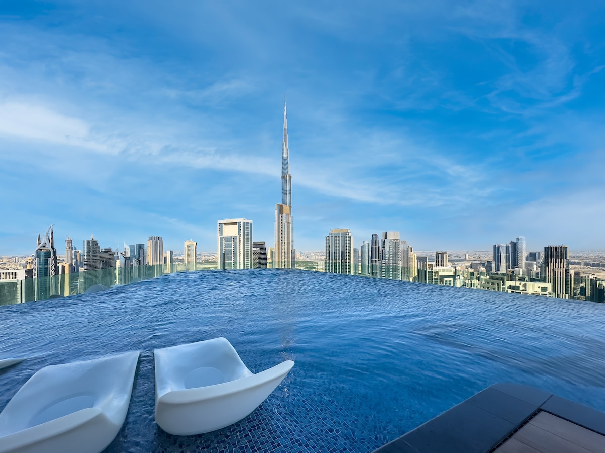 An infinity pool is featured on the rooftop, offering panoramic views of the skyline, including the Burj Khalifa. The tranquil water blends seamlessly into the horizon under a clear blue sky, while two sun loungers are positioned at poolside for relaxation.