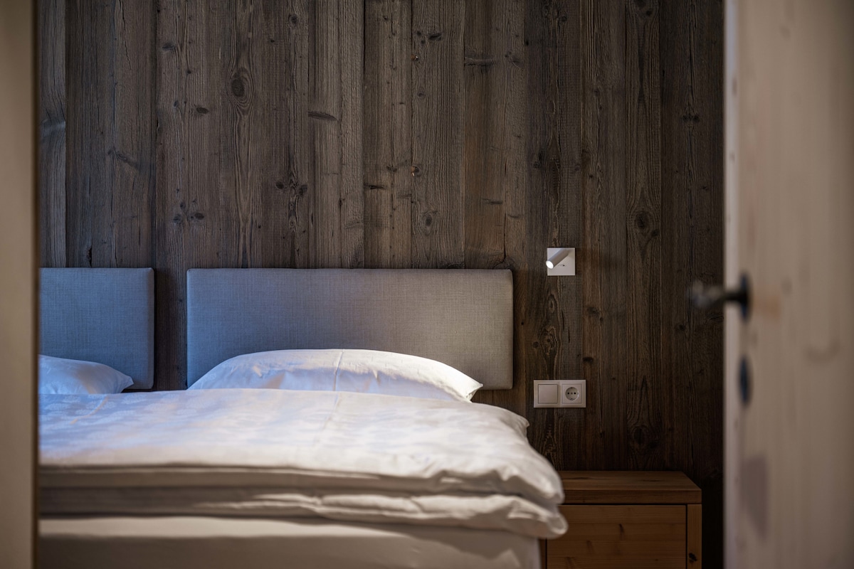 A cozy bedroom is featured, showcasing a king-size bed with soft linens and a simple headboard against a wooden wall. Minimalist side tables are positioned on either side, illuminated by subtle wall-mounted lights.