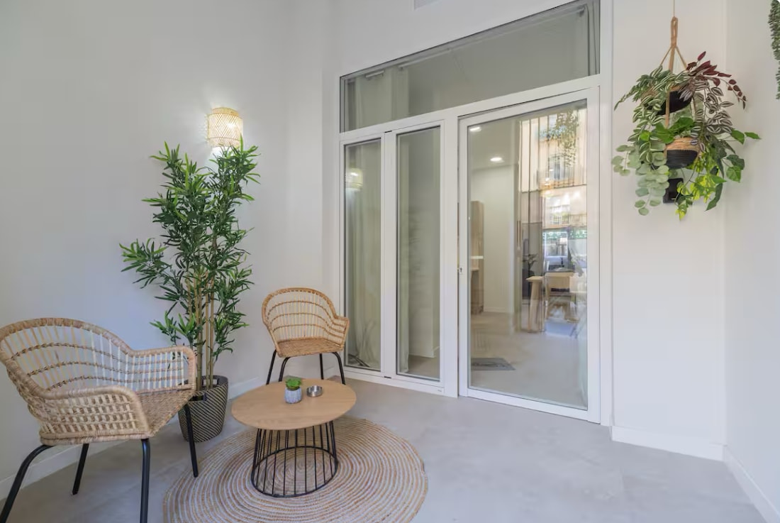 A light-filled corner of the studio features two woven chairs positioned around a round wooden table. A soft area rug lies beneath, with a small plant on the table. Large glass doors allow natural light to fill the space, creating a sense of openness.