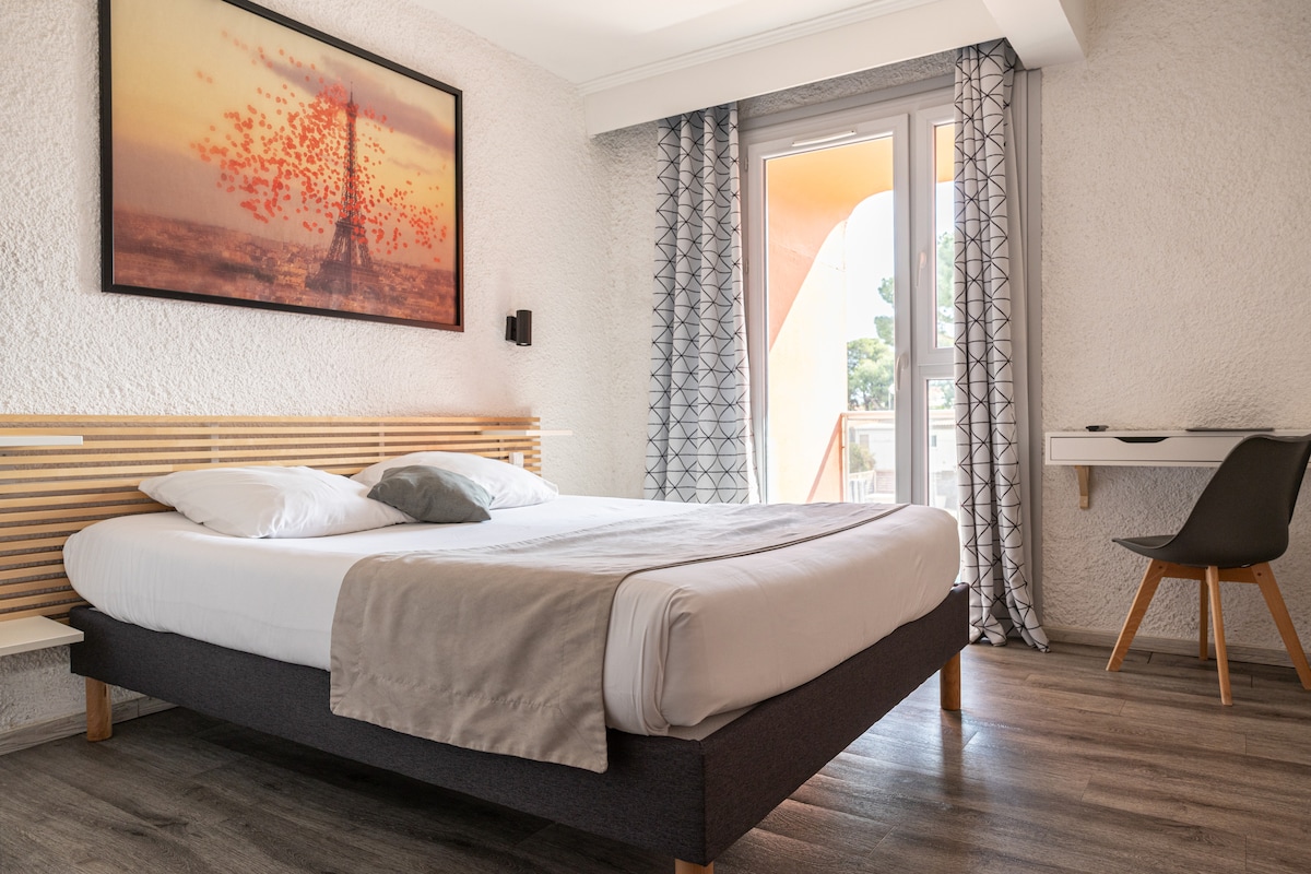 A bright and airy guest room features a spacious bed with light bedding and a soft throw blanket. A large window allows natural light to fill the space, showcasing a wall-mounted artwork of a sunset with the Eiffel Tower. A small desk and chair are positioned to the side.
