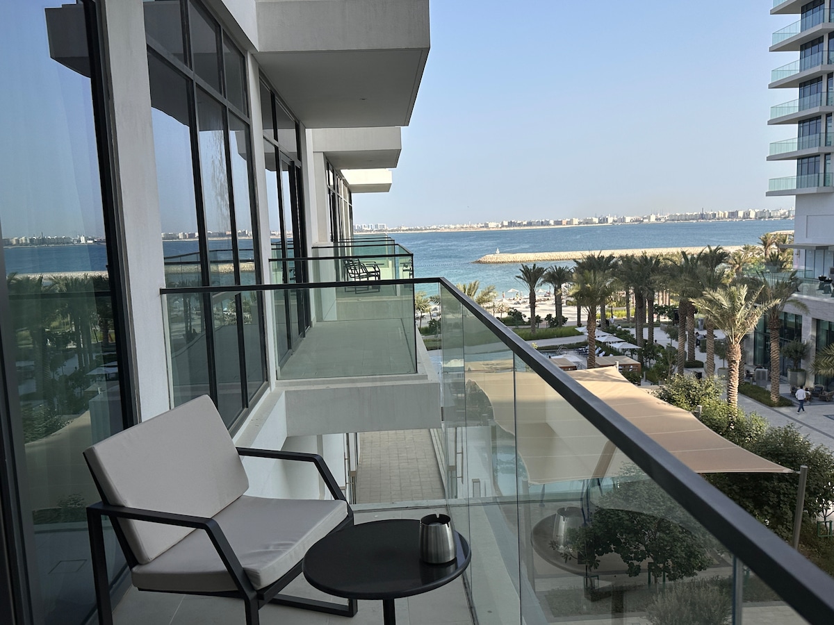 A serene balcony offers a glimpse of the stunning sea view and palm trees below. A small table sits alongside a single chair, creating a peaceful spot to relax. The glass railing allows for unobstructed sightlines of the beachfront and nearby amenities.