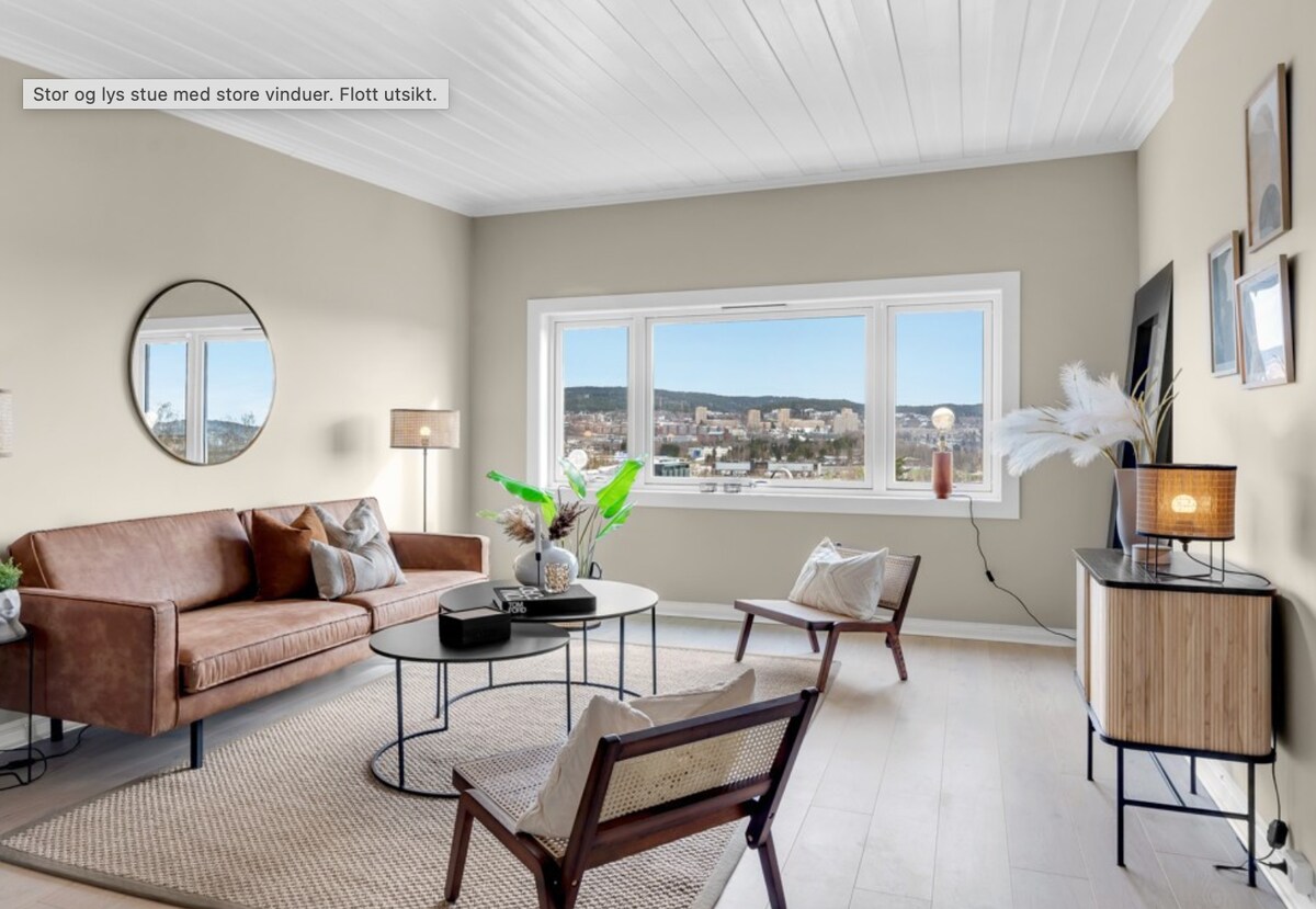 A spacious living room features large windows that provide a city view. A brown leather sofa is paired with two chairs and a round coffee table. A circular mirror and framed artwork adorn the walls. Natural light brightens the area, enhancing the modern decor.