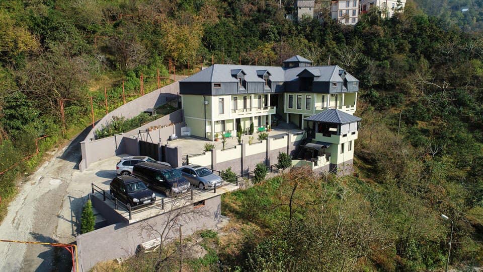 An aerial view captures the unique architecture of the property, characterized by multiple levels and a modern design. Surrounded by greenery, the structure features a well-maintained driveway with parked vehicles, leading to inviting outdoor spaces and a serene hillside environment.
