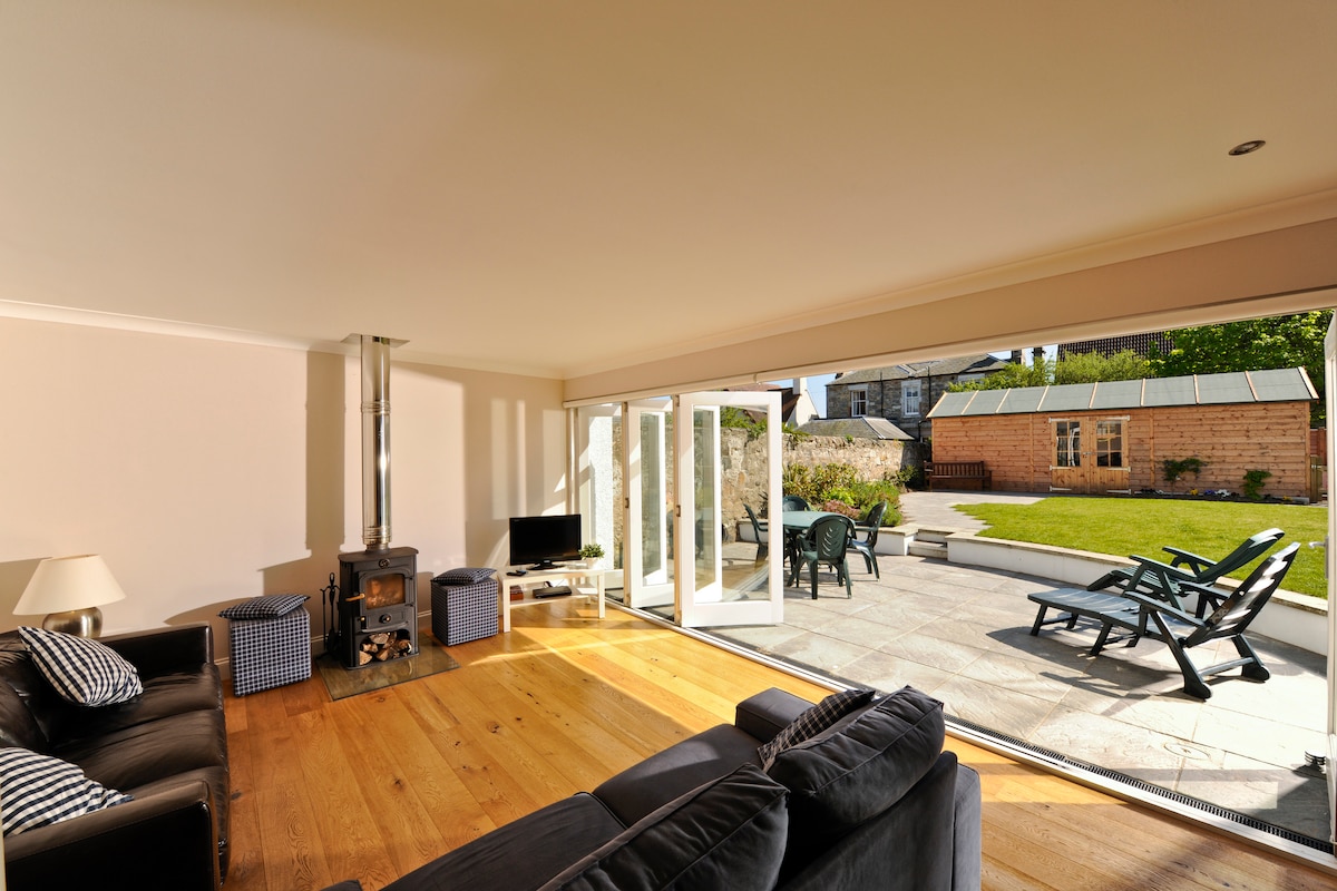 The open plan living area features polished wooden floors and a cozy wood burning stove. Expansive bi-fold doors open onto a private, sunny garden, showcasing outdoor seating options. A television is positioned nearby, contributing to the contemporary yet comfortable ambience.