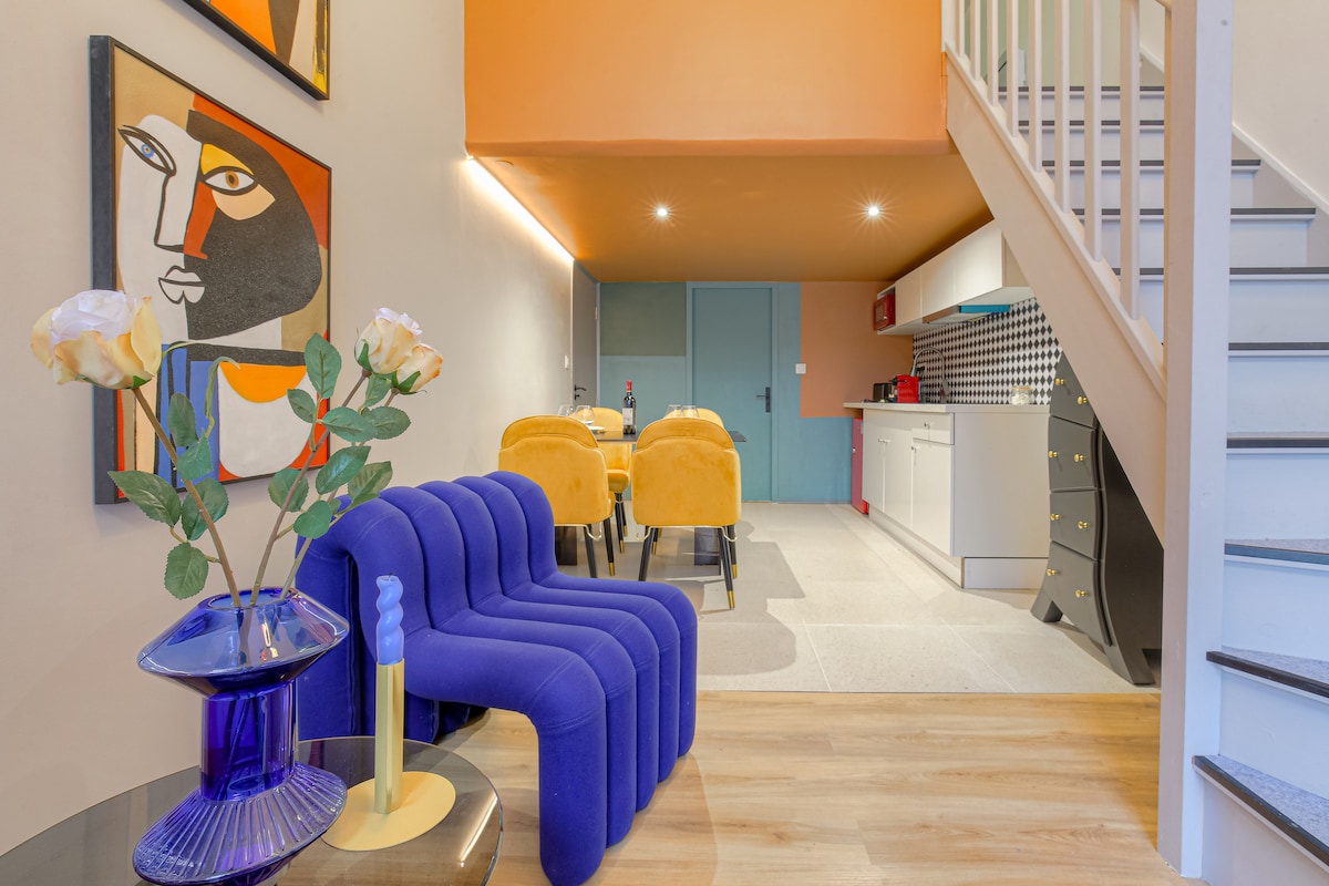 A modern living space features a colorful art piece alongside a blue accent chair and a transparent vase with flowers. A dining table with yellow chairs is visible in the background, complemented by a kitchen equipped with vibrant red cabinetry and black-and-white tiled backsplash.