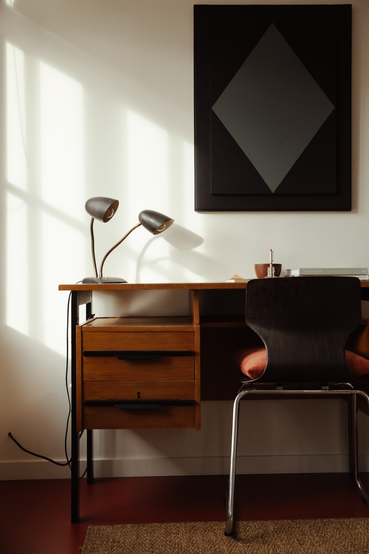 A well-lit workspace features a wooden desk equipped with two drawers. A lamp with two adjustable arms illuminates the area, complemented by a black chair with a red cushion. A large framed artwork is displayed above the desk, casting intriguing shadows on the wall.