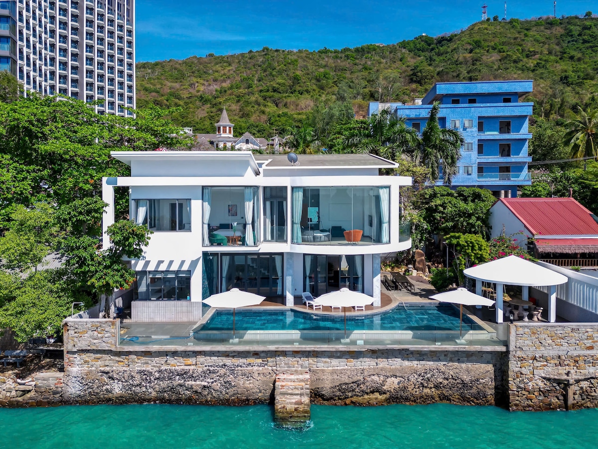 The villa is seen from an elevated perspective, showcasing its modern architecture with large glass windows. A terrace features sun loungers beside an infinity pool that overlooks the ocean. Surrounding greenery and the rocky shoreline are visible, enhancing the coastal setting.