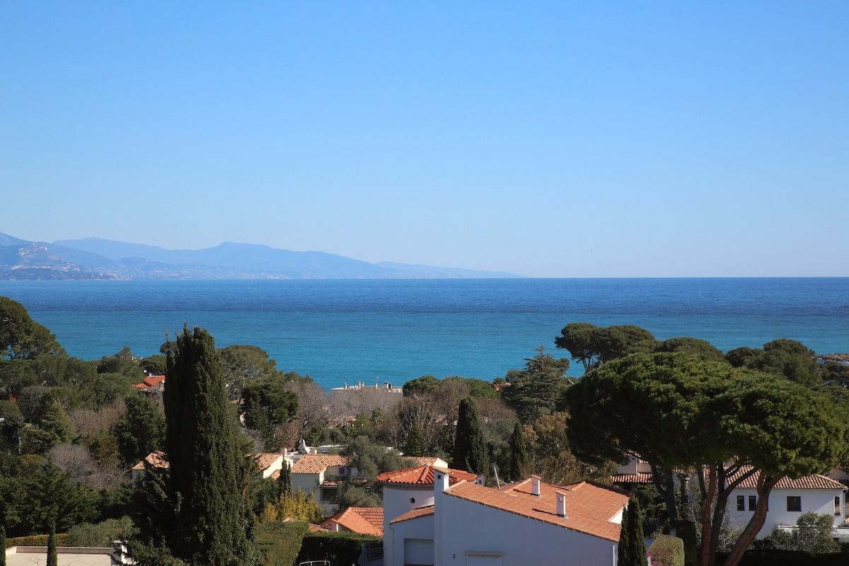 A panoramic view of the coastline is presented, showcasing tranquil blue waters meeting clear skies. Verdant trees and terracotta-roofed houses can be seen in the foreground, adding contrast to the scenic landscape. The mountains create a distant backdrop, enhancing the natural beauty of the area.