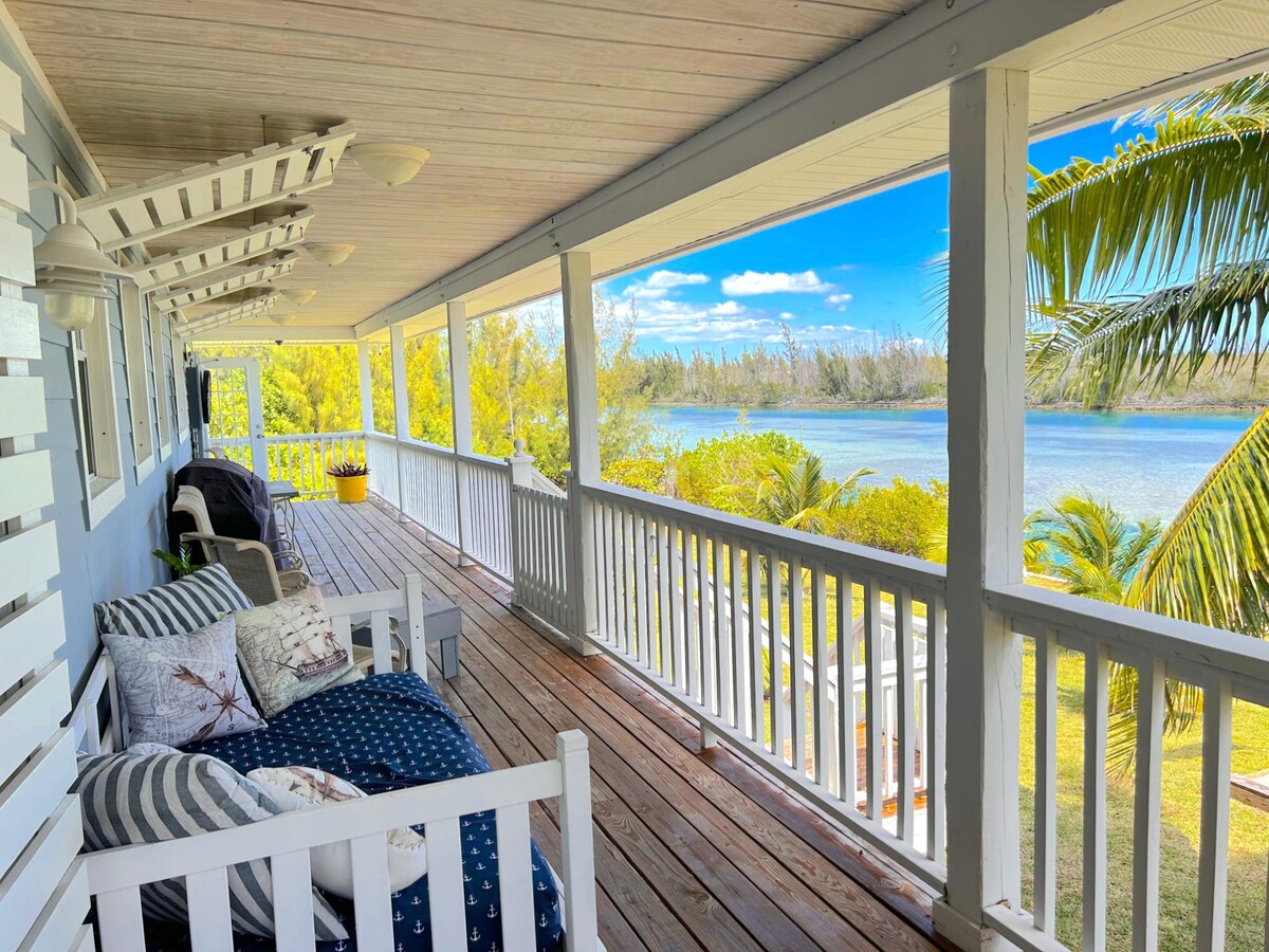 A covered porch with wooden flooring offers a serene view of the waterfront, framed by tropical trees. A white railing lines the area, while cushioned seating is positioned for relaxation. Sunlight brightens the space, emphasizing the tranquil surroundings and inviting atmosphere.