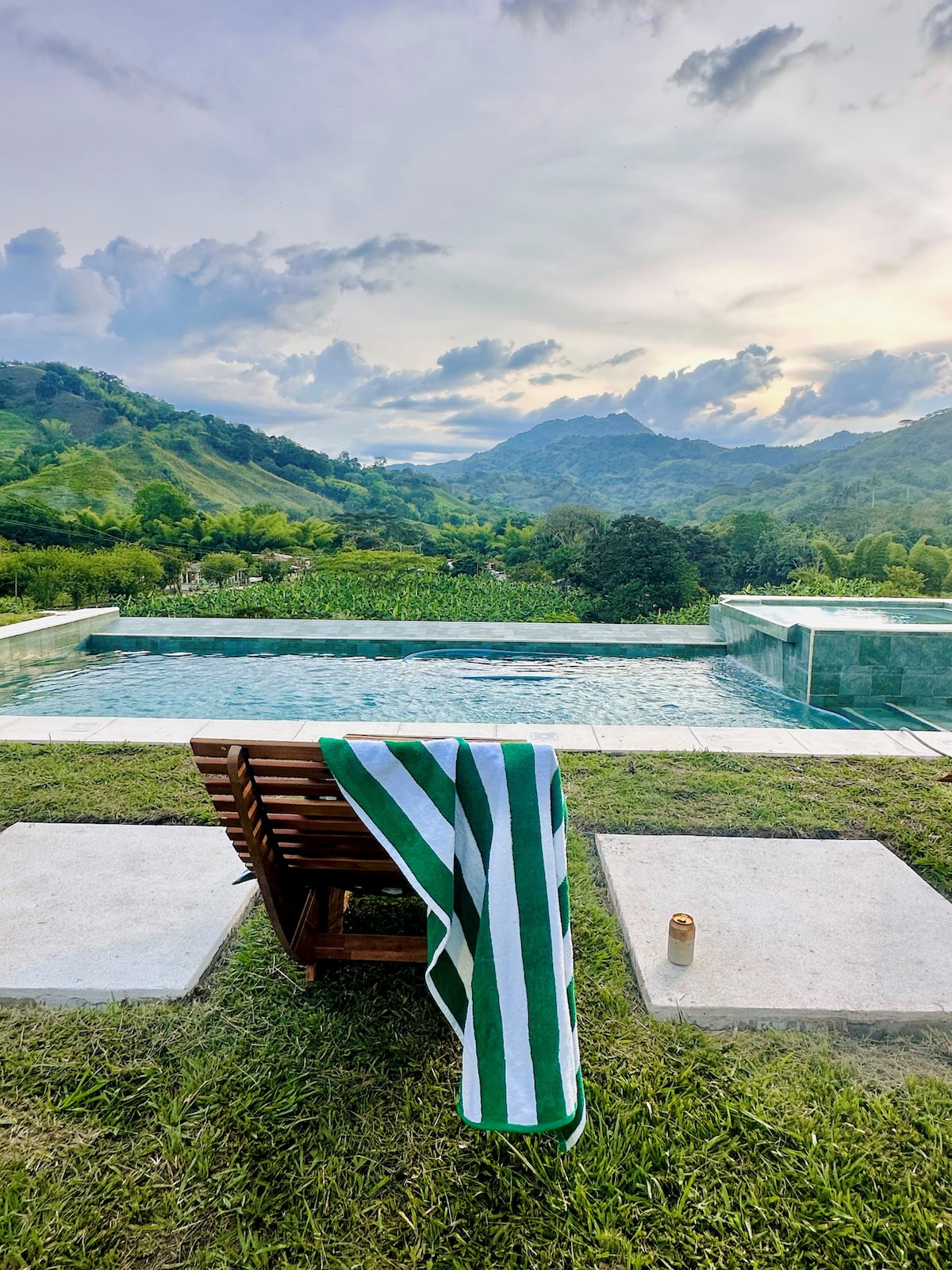 A wooden lounge chair is placed beside a pool, showcasing a striped green and white towel draped over it. The backdrop features expansive green hills and mountains under a partly cloudy sky, creating a serene outdoor setting.