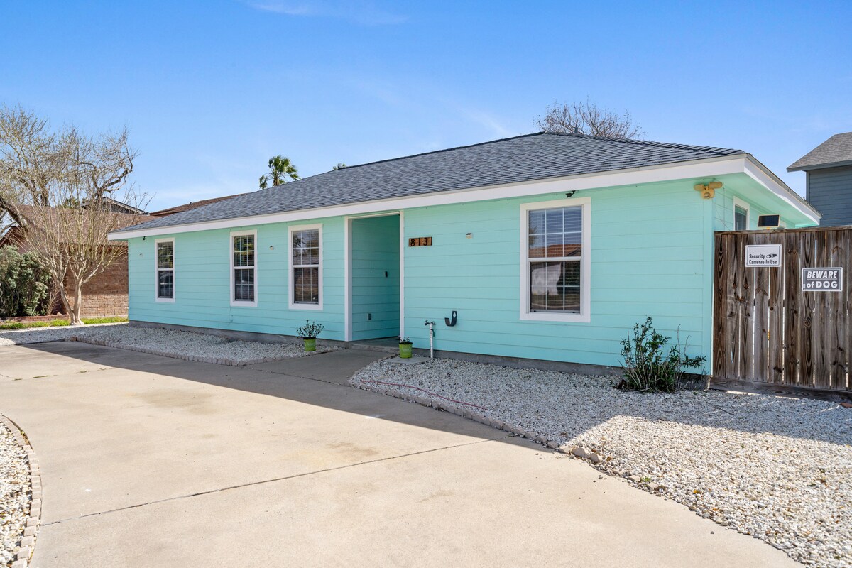A light blue coastal home is shown, featuring a welcoming facade with multiple windows and a gravel landscaping. The entrance is visible from a concrete driveway, surrounded by greenery and features a wooden fence for added privacy.