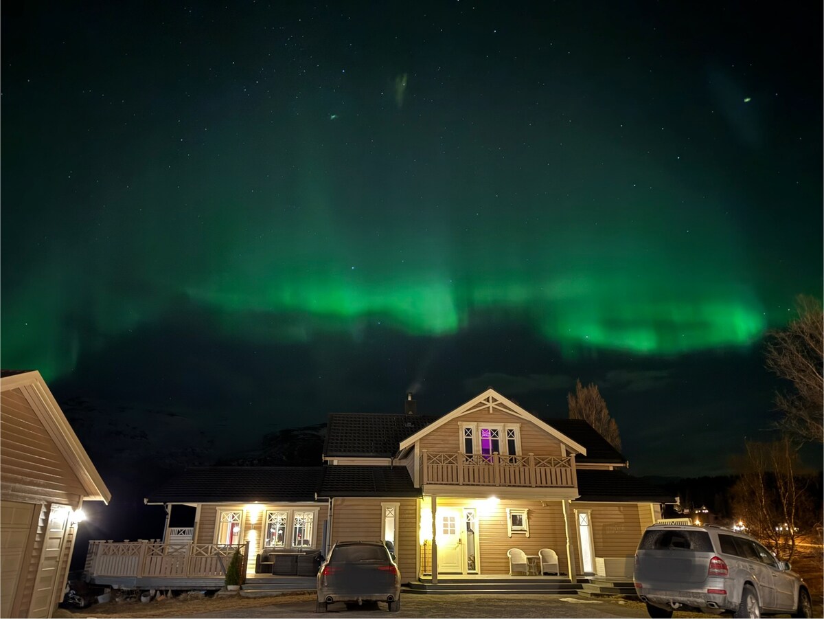 A charming house is illuminated under a vibrant display of green auroras. The two-story structure features a balcony, visible windows, and outdoor seating. Several cars are parked nearby, surrounded by a clear night sky filled with stars.