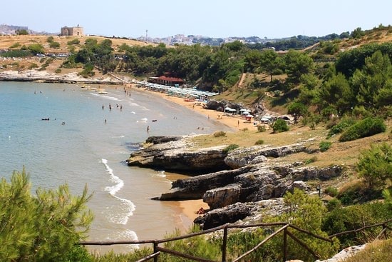 The image depicts a sandy beach with gentle waves lapping at the shore. Sunbathers can be seen scattered along the coastline, while lush green hills rise in the background. Sun loungers and umbrellas are visible, providing shaded areas for relaxation.