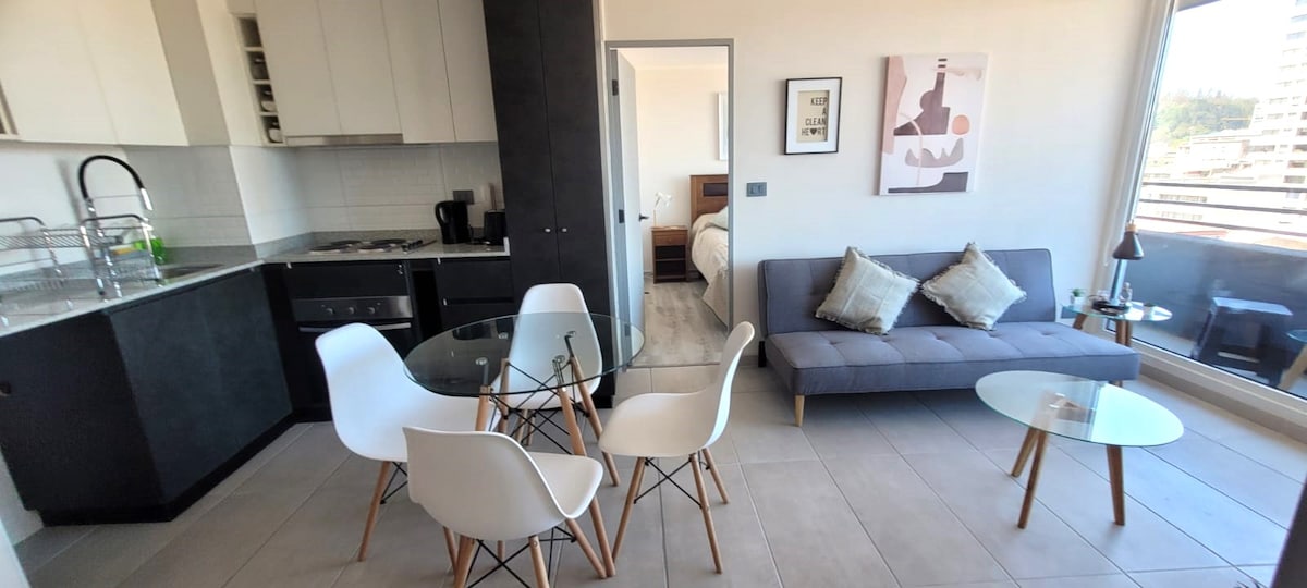 A modern living space is presented with a glass dining table surrounded by four white chairs. A gray sofa is positioned adjacent to a small coffee table. The kitchen area features sleek white cabinetry and stainless steel appliances. Large windows provide natural light and city views.