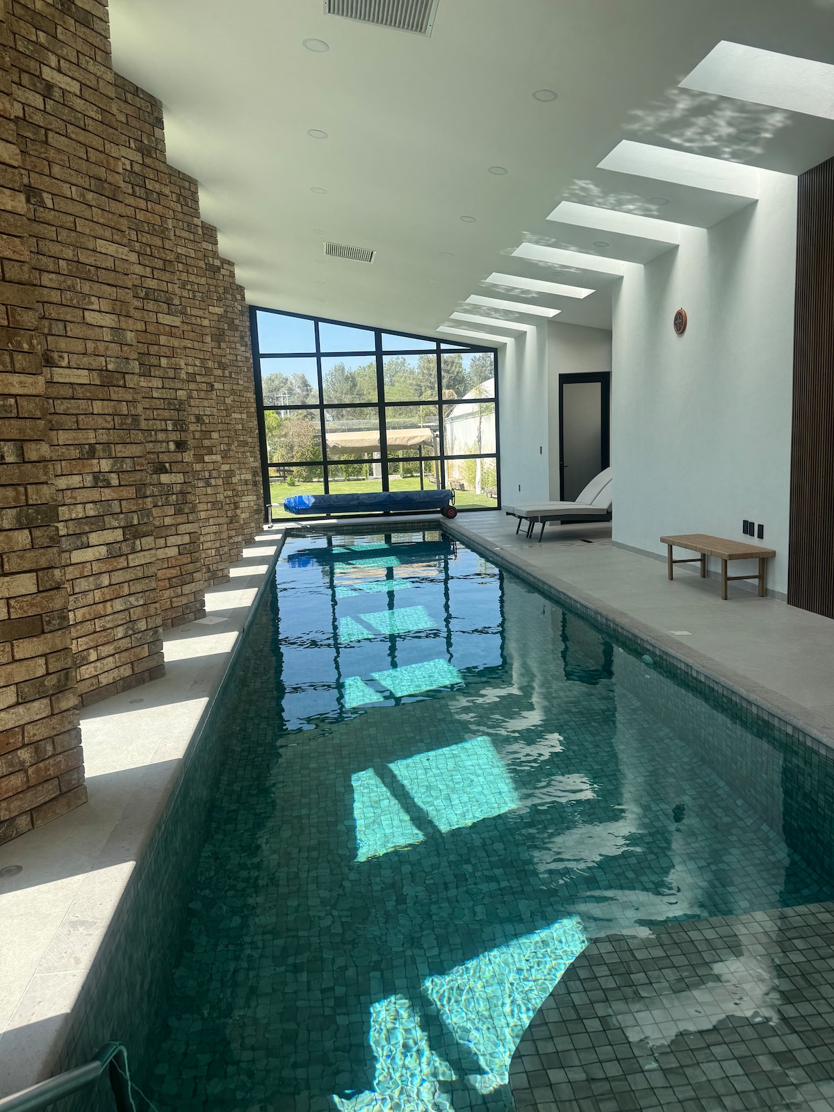 An indoor swimming pool is presented, with sunlight filtering through large windows above. The water reflects light, creating a serene ambience. A stone wall provides texture, while minimalist lounge chairs are positioned beside the pool for relaxation.