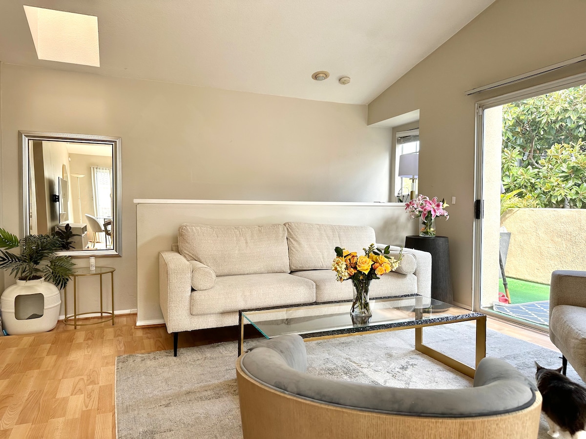 A light and airy living area features a soft beige sofa, a glass coffee table, and two elegantly upholstered chairs. Natural light enters through a sliding glass door, which opens to a patio adorned with greenery. A mirror and decorative flowers add subtle accents to the space.
