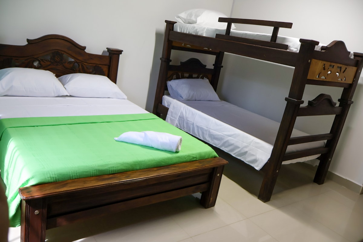 A well-lit sleeping area features a spacious bed covered with a green blanket, accompanied by a bunk bed in dark wood. Fresh linens are neatly arranged on both beds, creating an inviting space for relaxation.