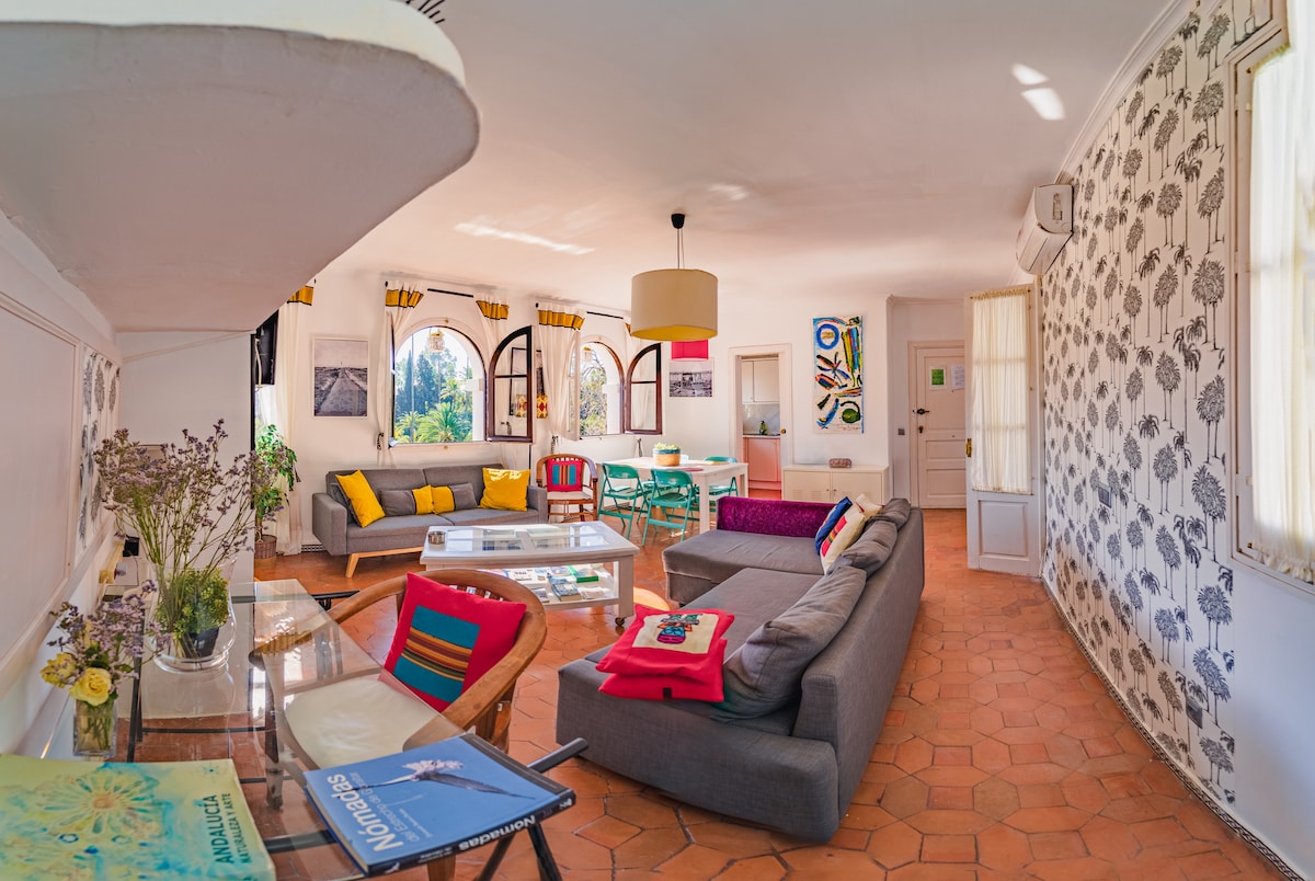 A spacious living area features multiple seating arrangements, including sofas and armchairs, complemented by decorative cushions. Large windows allow natural light to fill the room, while colorful artwork and vibrant décor enhance the inviting atmosphere.