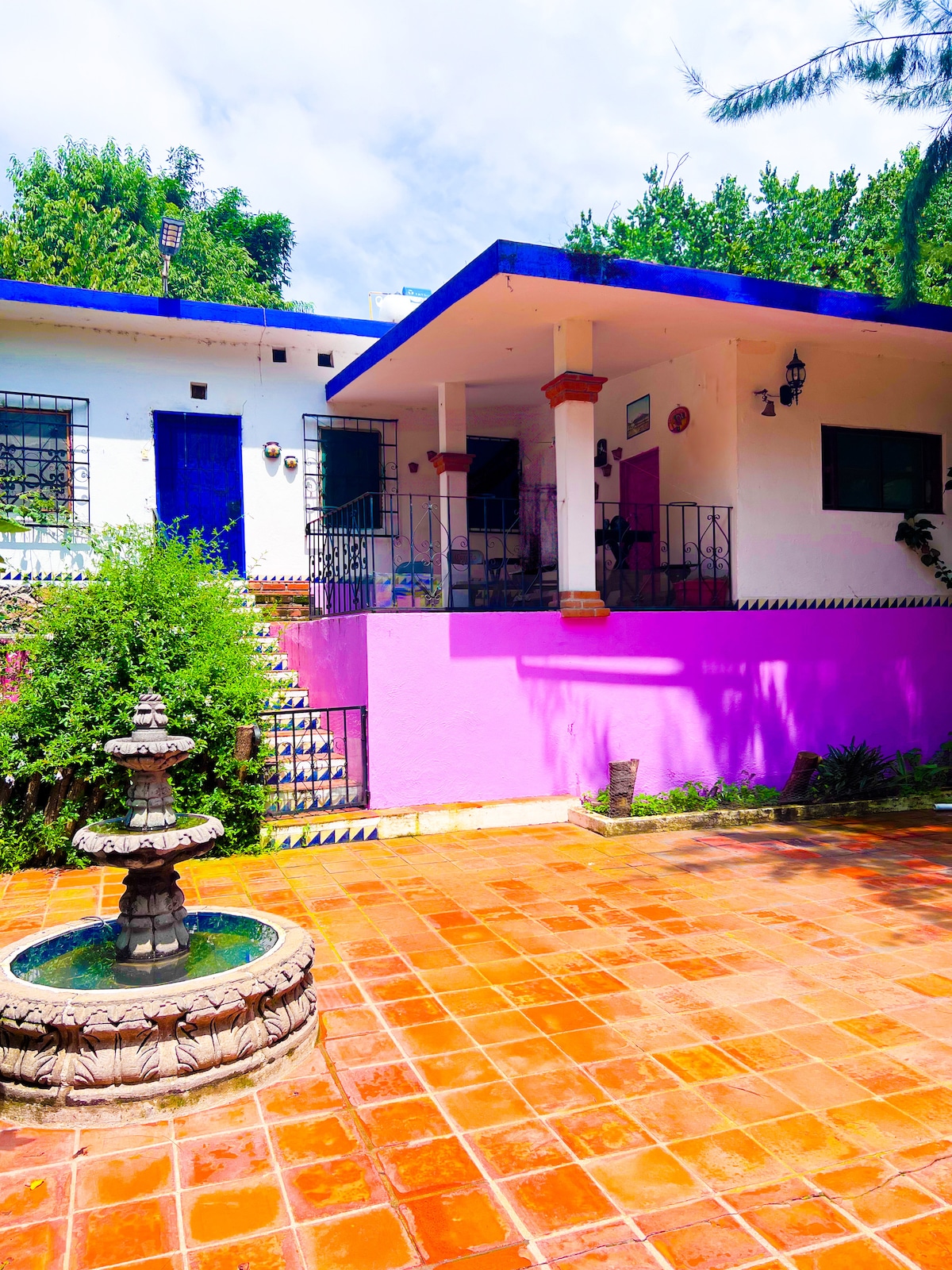 A rustic house is presented with a vibrant color scheme, featuring a blue roof and pink walls. A decorative stone fountain is situated in the foreground, surrounded by terracotta tiles, and lush greenery is visible in the background, adding a lively touch to the outdoor space.