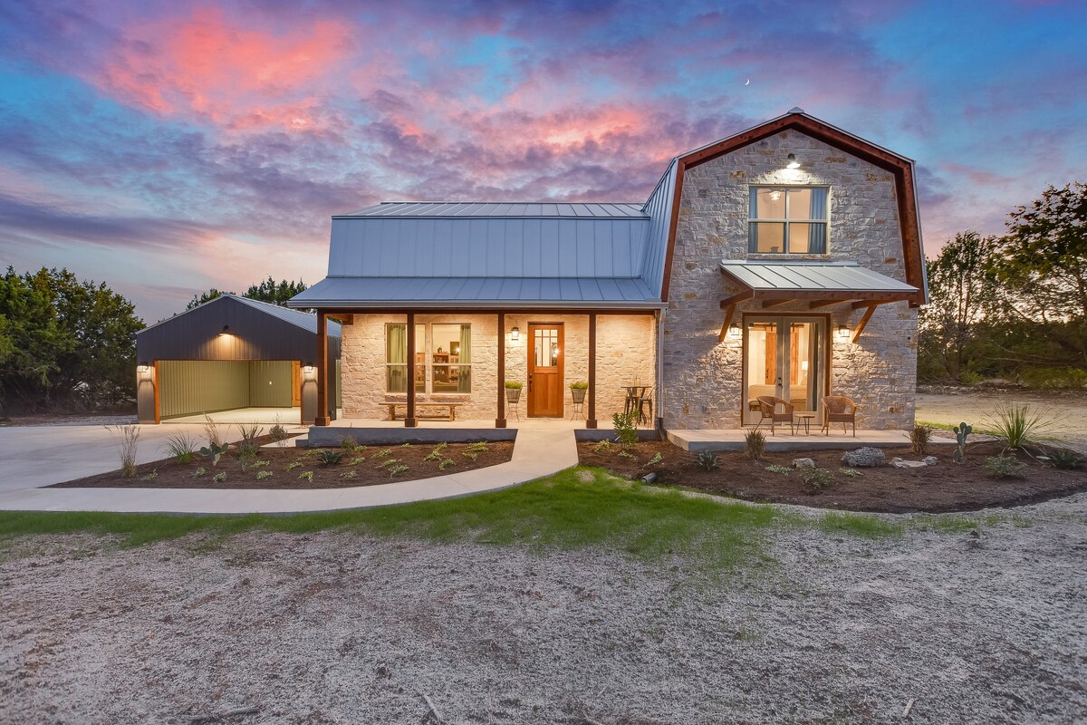 A modern farmhouse exterior is showcased against a vibrant evening sky. The structure features a combination of stone and metal elements, with large windows allowing for natural light. A well-maintained yard is visible, complete with carefully arranged landscaping and pathways leading to the entrance.