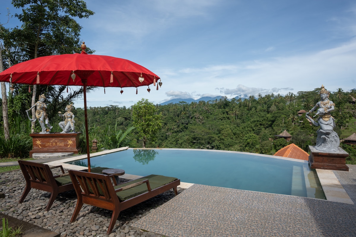 A private swimming pool is surrounded by natural greenery, with two lounge chairs positioned beside it. A vibrant red umbrella provides shade. Two decorative statues are situated nearby, overlooking the landscape, which features lush hills and distant mountains.
