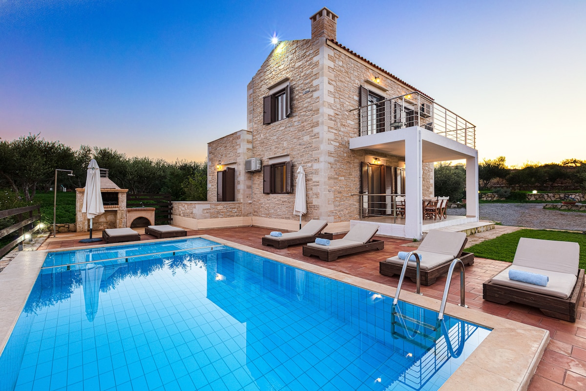 The villa's exterior features a private swimming pool bordered by sun loungers and umbrellas. A stone barbecue area is visible nearby, alongside a shaded dining space. The two-story structure showcases a combination of stone and modern design, set against a backdrop of olive trees at dusk.