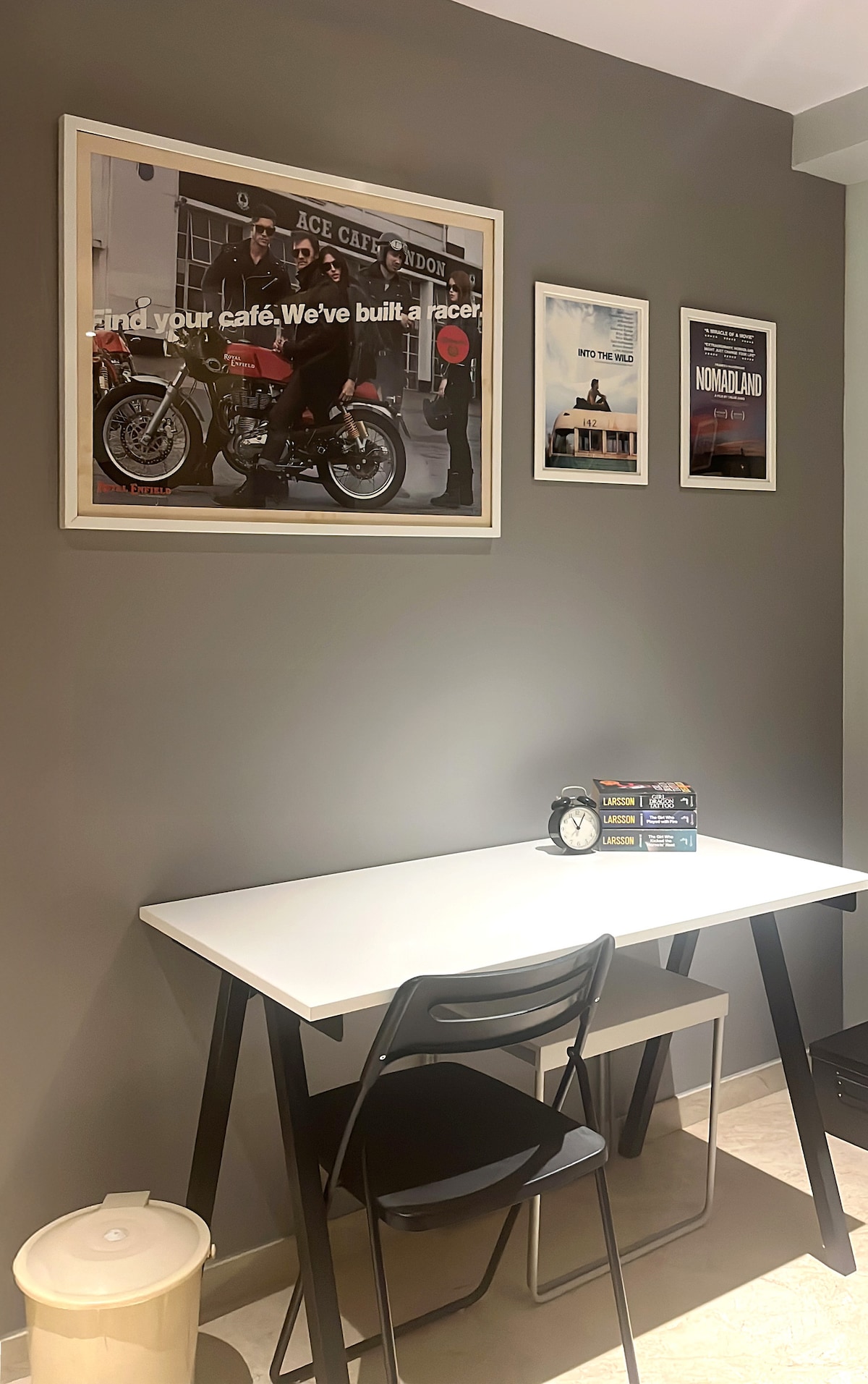 A modern workspace features a sleek white table accompanied by a black chair. A framed poster of motorcycles adorns the grey wall, alongside three art prints that add character. A clock and a stack of books sit atop the table, while a wastebasket is positioned nearby.