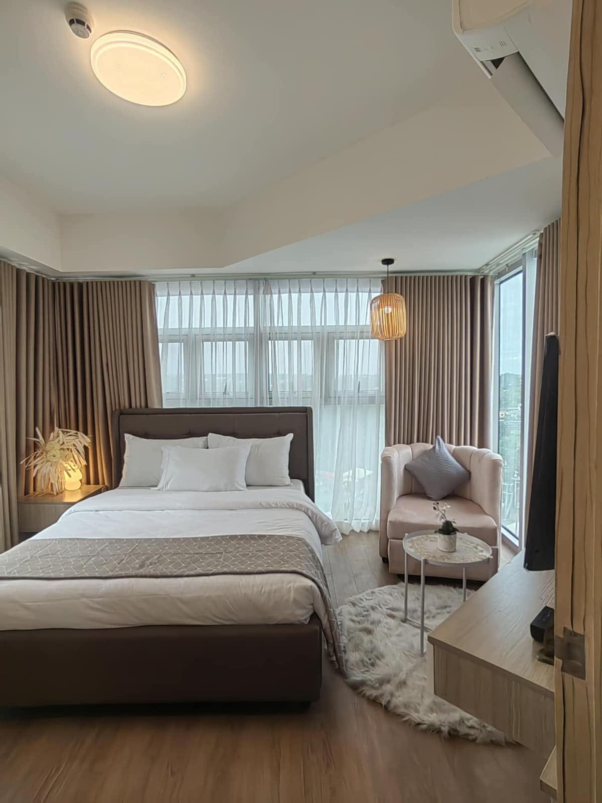 A bright bedroom features large windows draped with light curtains, allowing natural light to fill the space. A spacious bed is positioned against a backdrop of soft, neutral tones. A cozy armchair is placed beside a small side table, creating a relaxing corner. Warm lighting is provided by a pendant lamp.