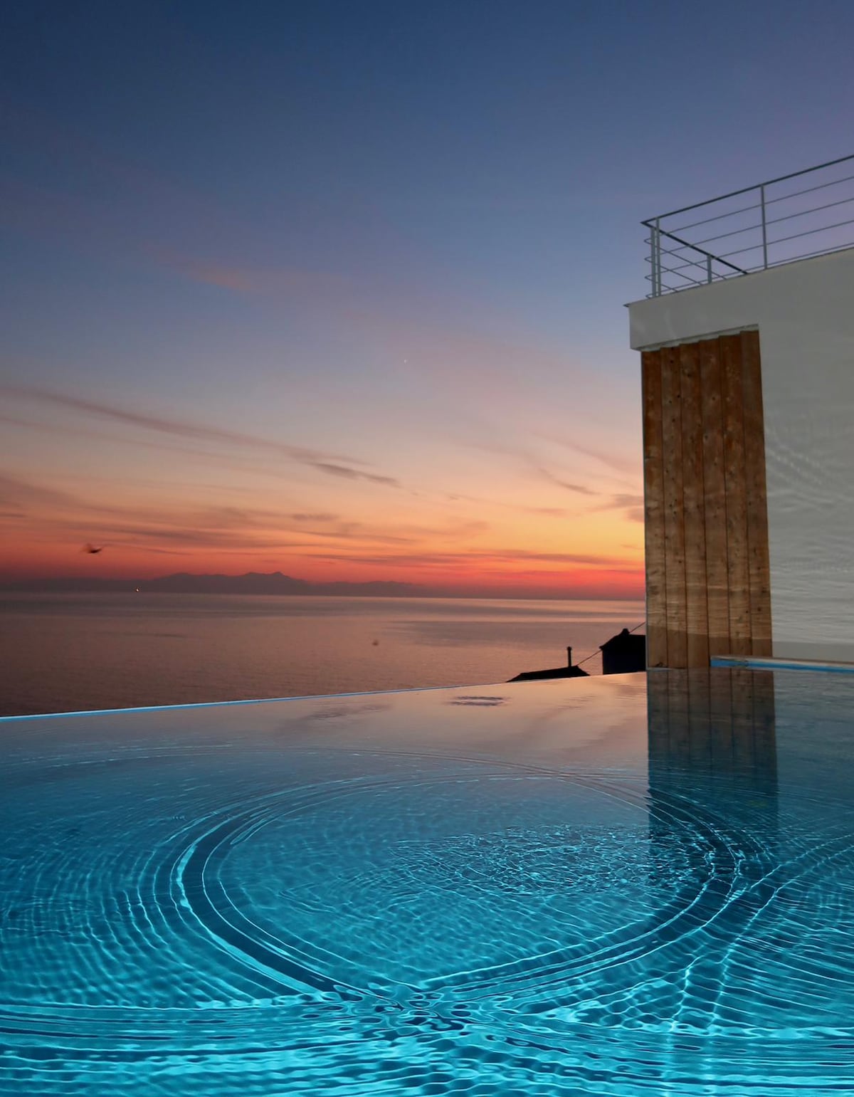 The image captures an infinity pool featuring shimmering water, reflecting the vibrant colors of a sunset. The serene landscape overlooks the sea, with the distant horizon merging into soft hues of orange and purple. The pool edge is framed by a modern building structure.